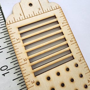 May include: A wooden bookmark with a scalloped top and a series of rectangular cutouts. The bookmark features inch and centimeter markings along the edges, with small holes near the bottom. The design is made of light-colored wood.