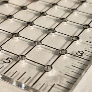 May include: A clear acrylic quilting ruler with black grid lines and measurement markings. The ruler features a grid pattern with evenly spaced holes, and the numbers 5 and 8 are visible on the edge. Ideal for sewing and quilting projects.