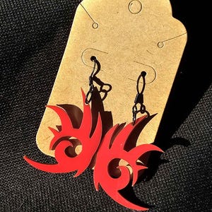 May include: A pair of red and black earrings featuring a stylised flame design. The earrings are hanging from a brown card.