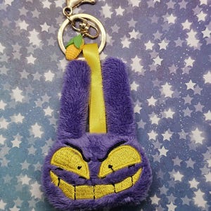 Può includere: Un portachiavi in ​​peluche viola con un viso giallo ricamato e un sorriso malvagio. Il portachiavi ha un fermaglio color oro, un nastro giallo e un piccolo ciondolo a forma di mais.
