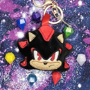 May include: A black and red plush keychain featuring Shadow the Hedgehog from the Sonic the Hedgehog video game series. The keychain is surrounded by colorful gems.