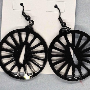 May include: A pair of black, circular earrings. Each earring features a wheel-like design with radial spokes and a central, elongated cutout. The earrings hang from black ear wires and are displayed on a white card.