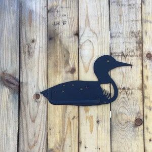 May include: Black metal silhouette of a loon bird mounted on a wooden wall. The loon is facing left and has a long beak. The wall is made of vertical wooden planks.