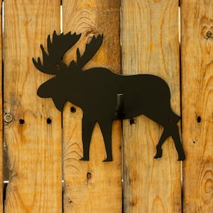 May include: Black metal moose silhouette wall hook mounted on a wooden fence.