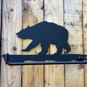 May include: Black metal bear silhouette towel rack. The bear is walking to the right and the towel rack is attached to a wooden wall.