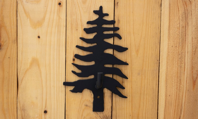 May include: Black metal pine tree wall hook with a single hook on the bottom. The hook is mounted on a light wood background.
