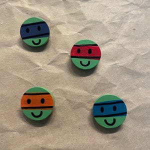 May include: Four green, round wooden Teenage Mutant Ninja Turtles-themed magnets. Each magnet features a different colored mask: blue, red, orange, and blue. Each has a smiling face.