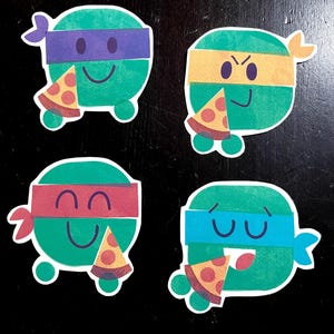 May include: Four cartoon stickers featuring green turtle-like characters with different colored headbands and expressions. Each character holds a slice of pizza. The headbands are purple, yellow, red, and blue. The stickers are on a black surface.