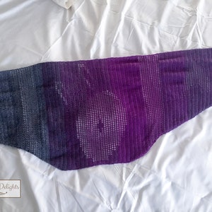 Puede incluir: Una bufanda de crochet de color morado y azul degradado con un patrón de encaje. La bufanda está doblada por la mitad y se encuentra sobre una superficie blanca.