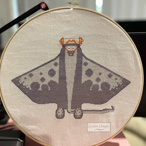 May include: A cross-stitch embroidery of a manta ray with a white and orange patterned head, set in a round wooden embroidery hoop. The embroidery is signed "Nerdyish Delights".