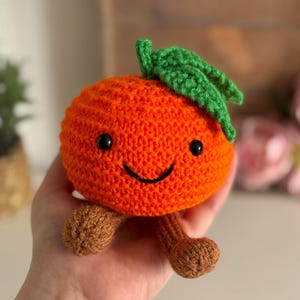 May include: A crocheted orange fruit toy with a smiling face, black eyes, and green leaves. The orange has brown legs and is held in a hand. The toy is made of orange yarn.