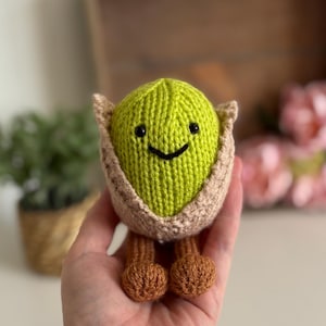 Könnte beinhalten: Ein handgestricktes Avocado-Plüschtier mit einem lächelnden Gesicht. Die Avocado ist grün, eingebettet in eine beige Schale, mit braunen Füßen. Das Spielzeug wird in einer Hand gehalten, mit einem verschwommenen Hintergrund aus Pflanzen und einer Holzkiste.