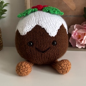 May include: A round, brown knitted plush toy shaped like a Christmas pudding. It features a white knitted icing top, green leaves, and a small red berry. The toy has two small brown feet, a stitched smile, and two black bead eyes on its front.