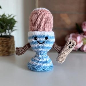 Può includere: Un giocattolo a forma di uovo lavorato a mano, con una parte superiore rosa, corpo a righe blu e bianche e braccia marroni. Tiene in mano una piccola fetta di pane tostato beige lavorata a maglia. Il giocattolo ha una faccia sorridente.