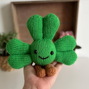 May include: A handmade green knitted creature with a clover-like design. The plush toy has a smiling face with black eyes and a mouth. It has brown feet and is held in a hand. The background includes a wooden box and a small plant.