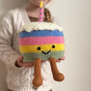 May include: A knitted birthday cake plush toy with a smiling face, rainbow stripes, and a white frosting top. The cake has brown legs and a candle with a yellow flame. The toy is held by a person.