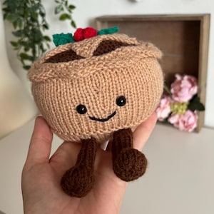 May include: A round, tan, knitted plush toy shaped like a pie with a dark brown interior, a smiling face with black bead eyes, and two dangling dark brown legs. The top of the pie features a decorative green leaf and two small red berries.