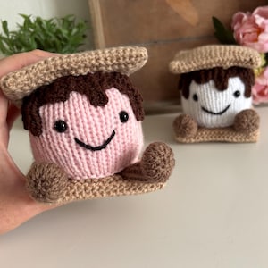 Könnte beinhalten: Zwei gehäkelte S'mores-Plüschfiguren mit einem hellrosa oder weißen Marshmallow-Kern, dunkelbraunen Schokoladentropfen und beigen Graham-Cracker-Stücken oben und unten. Jede Figur hat kleine schwarze Perlenaugen und ein gesticktes Lächeln.
