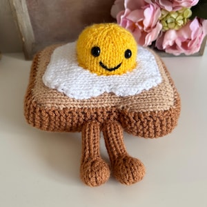 May include: A handmade, knitted toy resembling a piece of toast topped with a fried egg. The egg has a yellow yolk with a smiling face and a white egg white. The toast is tan and brown, with two brown legs. The toy is on a white surface.