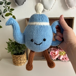 Puede incluir: Una tetera azul tejida con un ribete blanco y una cara sonriente. La tetera tiene patas marrones y un botón blanco en la parte superior. La tetera está sujeta por una mano. Hay corazones decorativos y plantas en el fondo.