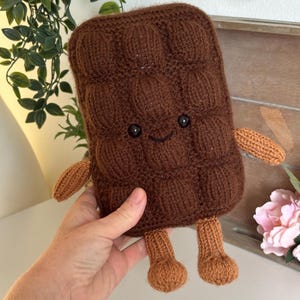 May include: A brown rectangular crochet plush toy shaped like a chocolate bar with a textured grid pattern. It features two small black bead eyes, a stitched smile, two light brown arms extending from the sides, and two light brown legs.
