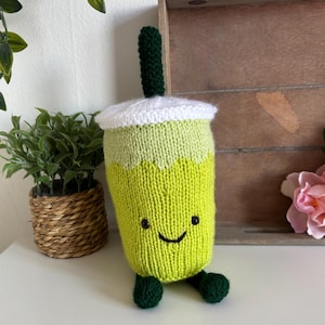 Op de afbeelding: Een limoengroen gebreid knuffeltje in de vorm van een boba-theebeker met een witte deksel, een donkergroen rietje en twee kleine donkergroene voetjes. De beker heeft een lachend gezichtje met twee zwarte kraaloogjes en een geborduurde zwarte mond.