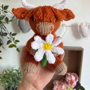 May include: A knitted stuffed animal shaped like a Highland cow with rust-coloured yarn, white horns, and a tan muzzle. The cow features a textured fringe of yarn on its head and is holding a large, crocheted white daisy with a yellow centre.