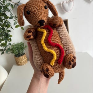 May include: A brown knitted dachshund toy shaped like a hot dog, featuring black plastic eyes, a black nose, and embroidered dark brown paw pads. The body is wrapped in a light tan knitted bun with wavy yellow mustard and red ketchup yarn lines.