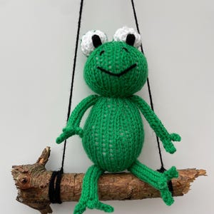 Puede incluir: Una muñeca de rana verde tejida a mano con ojos blancos y negros, sentada en un columpio de rama de madera. La rana tiene una cara sonriente y está suspendida por cuerdas negras, creando una pieza decorativa caprichosa.