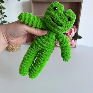 May include: A vibrant green crocheted frog toy with black eyes and a smiling mouth. The frog has outstretched arms and legs, made from soft, textured yarn. This plush toy is designed for children. The frog is a cheerful green colour.