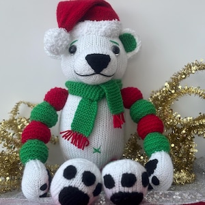 May include: A handmade knitted polar bear Christmas ornament. The bear is white with a red Santa hat, a green scarf, and red and green segmented arms. The bear has black eyes and a black nose. The bear's feet are white with black paw prints.