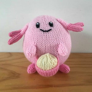 Puede incluir: Un peluche rosa tejido a mano con una cara sonriente, brazos pequeños y apéndices en forma de orejas. El juguete sostiene una bolsa de color amarillo claro. El peluche está hecho de hilo suave y se encuentra sobre una superficie de madera.