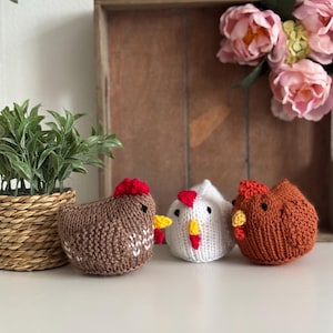 May include: Three knitted chicken figurines in brown, white, and orange, each with a red comb and yellow beak. The brown chicken has white accents. They are displayed on a white surface with a woven basket and faux flowers in the background.