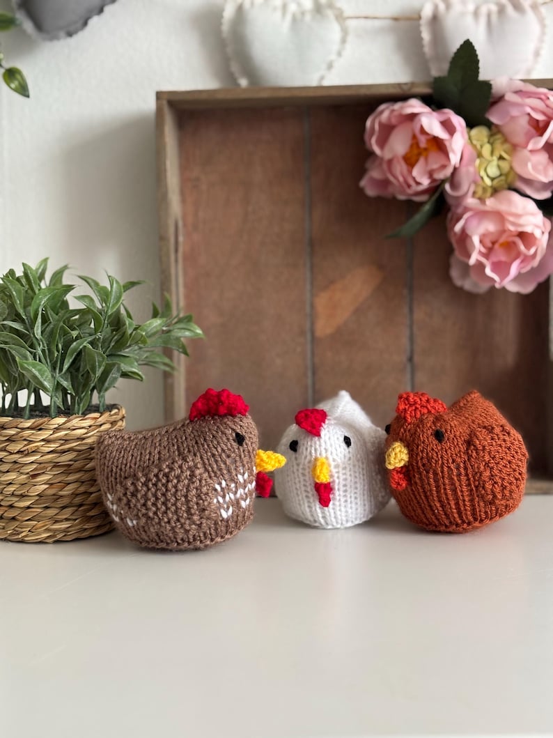 May include: Three knitted chicken figurines in brown, white, and orange, each with a red comb and yellow beak. The brown chicken has white accents. They are displayed on a white surface with a woven basket and faux flowers in the background.