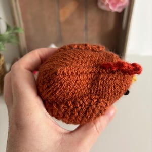 May include: A handmade, knitted, brown hedgehog toy with a red crest and yellow accents. The hedgehog is held in a hand, showcasing the detailed stitching and texture of the yarn. The toy is a charming, handcrafted item.