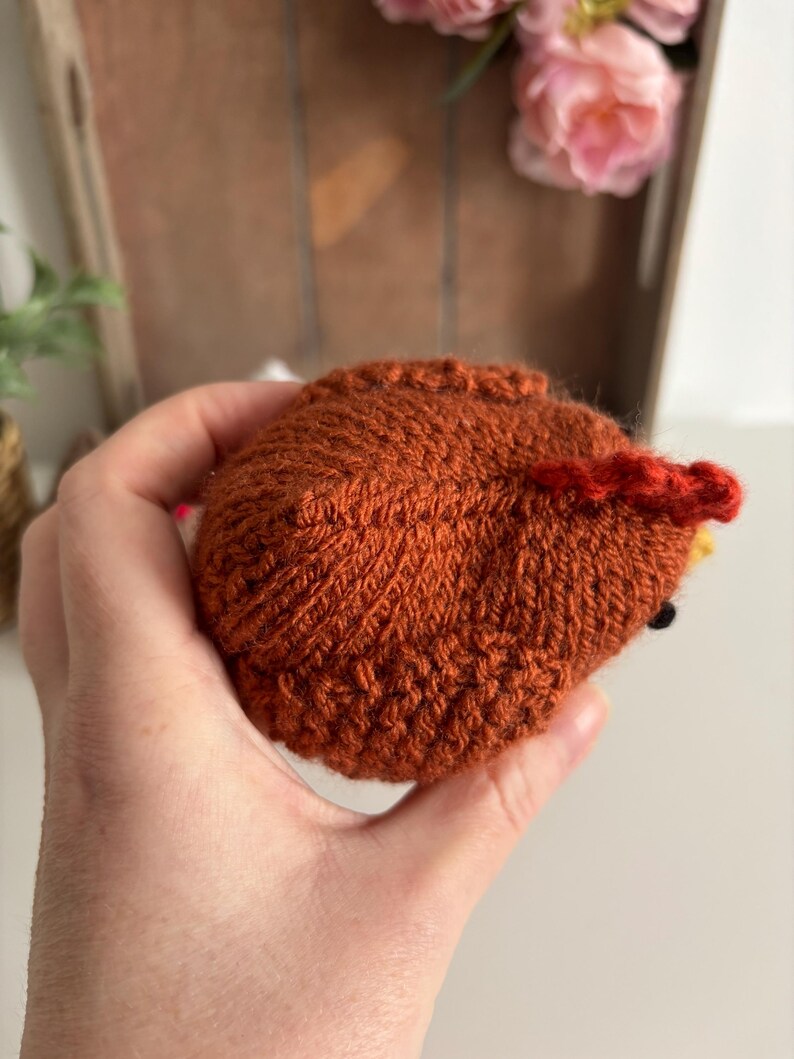 May include: A handmade, knitted, brown hedgehog toy with a red crest and yellow accents. The hedgehog is held in a hand, showcasing the detailed stitching and texture of the yarn. The toy is a charming, handcrafted item.