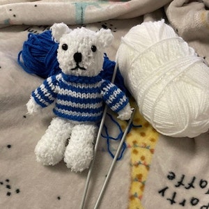 May include: A handmade white teddy bear wearing a blue and white striped sweater. The bear is surrounded by blue and white yarn, and knitting needles, suggesting a craft project. The bear has black eyes and a black nose.