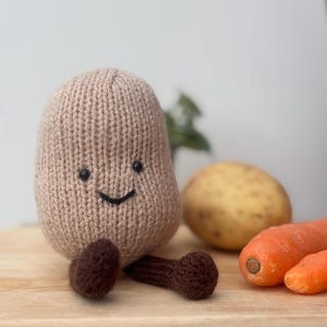 Puede incluir: Un peluche tejido con forma de patata sonriente en marrón claro con brazos y pies marrón oscuro. Una patata y dos zanahorias naranjas están junto al juguete sobre una superficie de madera. El juguete tiene ojos negros y una sonrisa cosida.