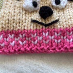 Pu&ograve; includere: Un cappello di lana con un viso di cartone animato. Il cappello &egrave; beige con un bordo rosa e bianco. Il viso ha occhi neri, un naso nero e una bocca nera.