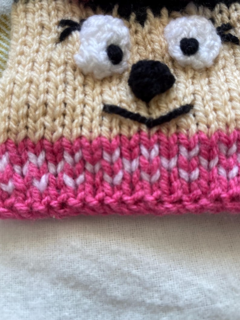 Pu&ograve; includere: Un cappello di lana con un viso di cartone animato. Il cappello &egrave; beige con un bordo rosa e bianco. Il viso ha occhi neri, un naso nero e una bocca nera.