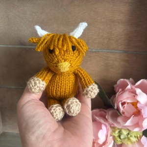 May include: A small, knitted Highland cow figurine. The cow is primarily mustard yellow with tan hooves and a tan muzzle. It has white horns and a fluffy head of hair. The cow is a handmade item.