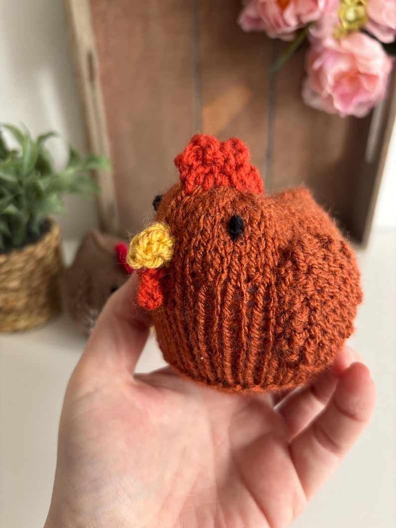 May include: A knitted chicken figurine in shades of brown, orange, and yellow. The chicken has a red comb and wattle, with black bead eyes. The small, round chicken is held in a hand, with a blurred background of plants and flowers.