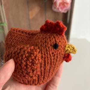May include: A hand-knitted, orange-brown chicken figurine with a red comb and wattle, and a yellow beak. The chicken has a black eye and is made of textured yarn. A charming, handmade decorative item.