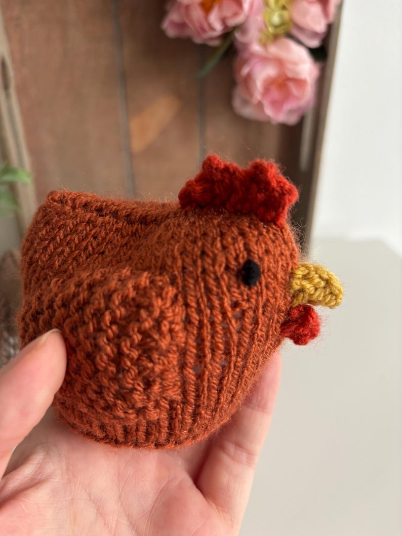 May include: A hand-knitted, orange-brown chicken figurine with a red comb and wattle, and a yellow beak. The chicken has a black eye and is made of textured yarn. A charming, handmade decorative item.