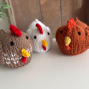 May include: Three knitted chicken figurines in brown, white, and orange. Each chicken has a red comb and yellow beak. The chickens are on a white surface with a wooden background.