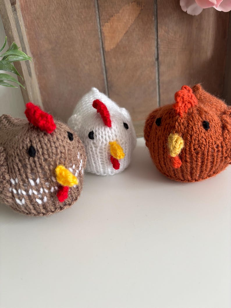 May include: Three knitted chicken figurines in brown, white, and orange. Each chicken has a red comb and yellow beak. The chickens are on a white surface with a wooden background.