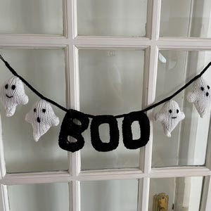 May include: A black knitted banner with the word "BOO" hanging on a white door with a string of white knitted ghosts.