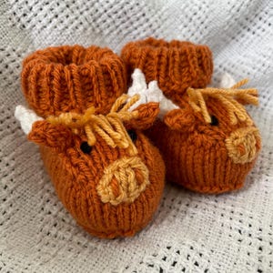 May include: A pair of knitted baby booties shaped like brown and white cows. The booties have a brown knit body with white accents for the horns and ears. The face is detailed with a brown nose and yellow eyes.