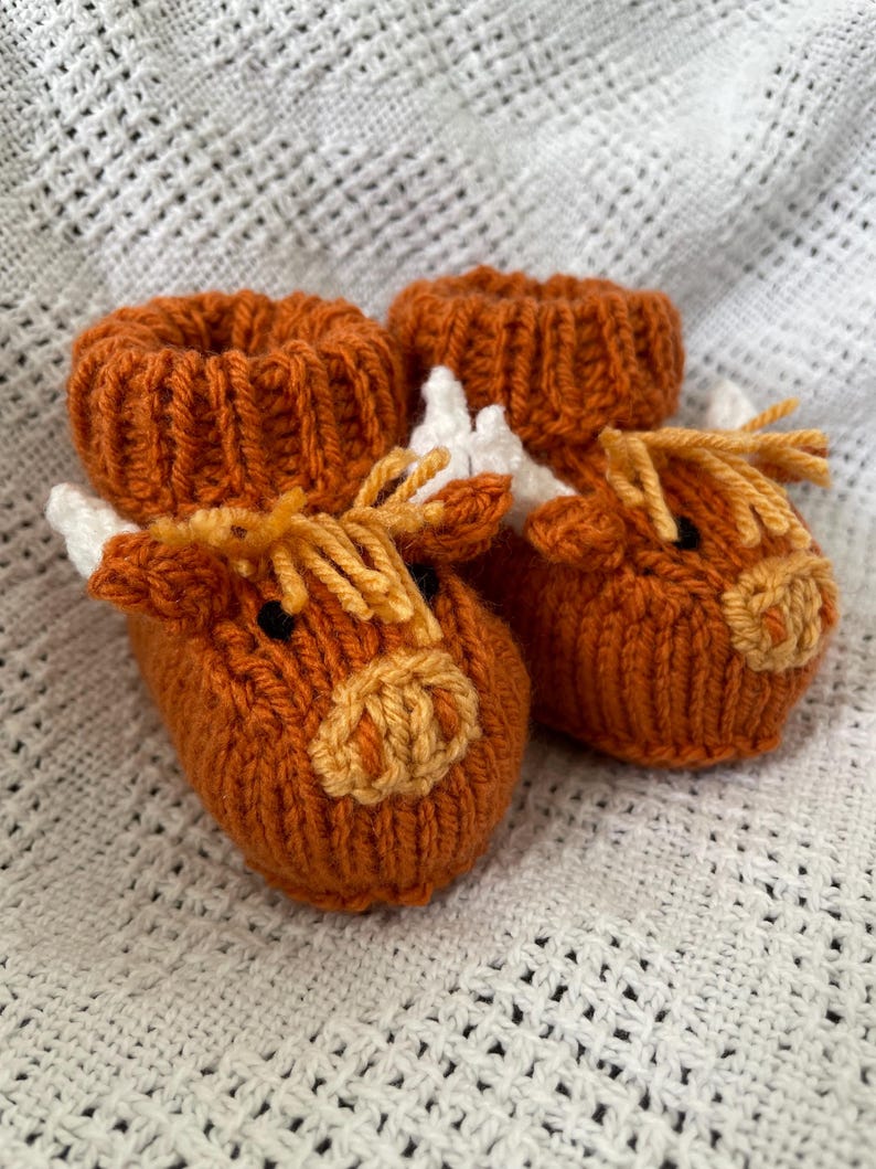 May include: A pair of knitted baby booties shaped like brown and white cows. The booties have a brown knit body with white accents for the horns and ears. The face is detailed with a brown nose and yellow eyes.