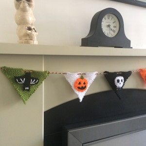May include: A knitted Halloween banner with green, white, orange, and black triangular flags. Each flag features a different Halloween icon: a bat, a pumpkin, a skull, and a ghost. The banner is hanging above a fireplace.
